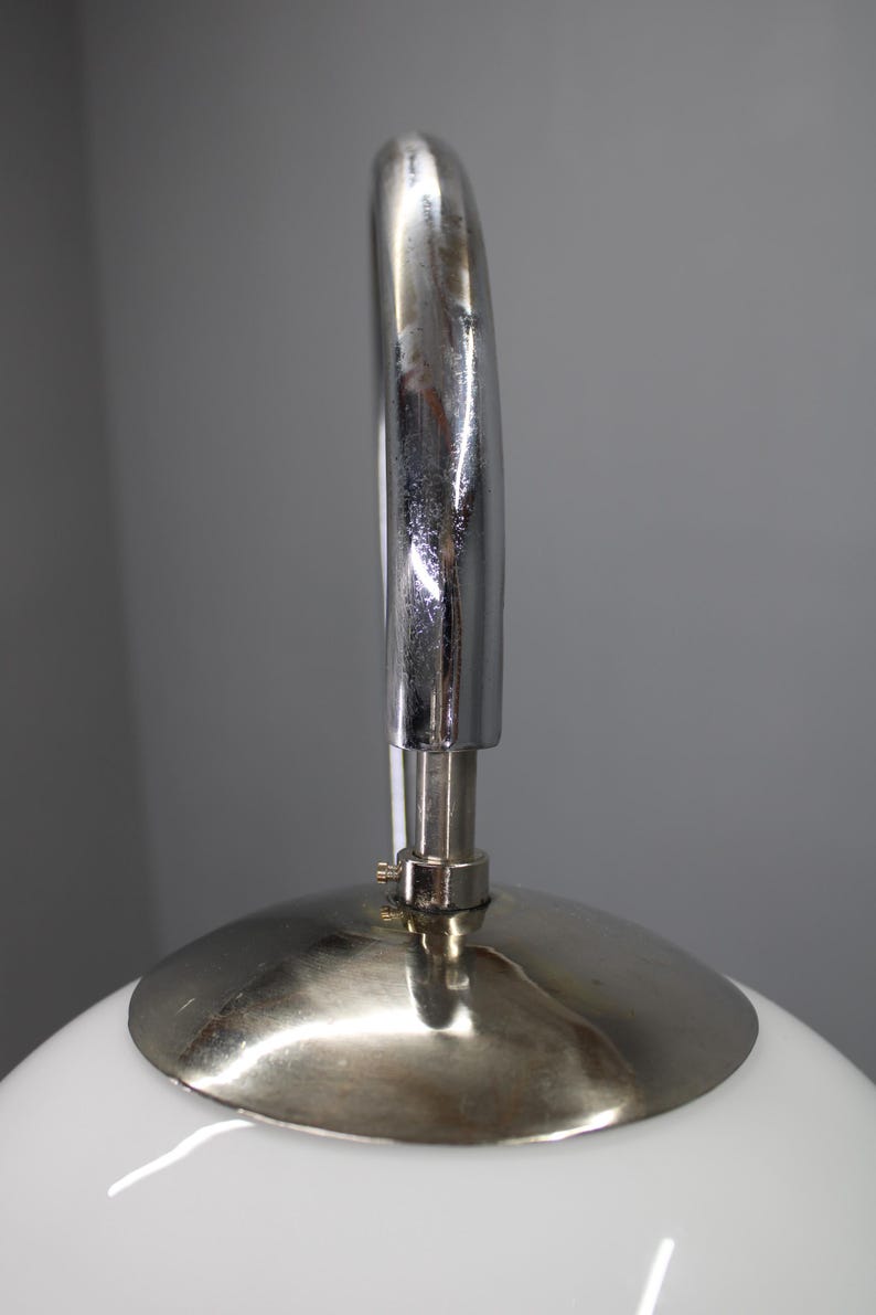 May include: Close-up of a white, rounded lamp with a silver metal handle and base. The handle arches over the top of the lamp, and the base is a circular disc. The lamp has a modern, minimalist design.