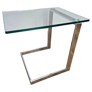 May include: A modern side table with a clear glass top and a polished metal frame. The table has a rectangular glass surface and a unique, angular base design. The metal frame is a bright silver color.