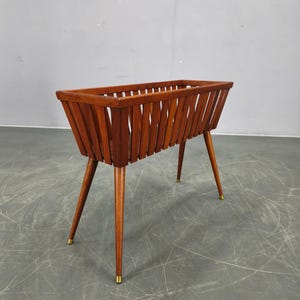 May include: A mid-century modern wooden planter with a rectangular basket design. The planter features angled wooden slats and slender, tapered legs with brass-colored tips. The wood has a warm, reddish-brown tone.
