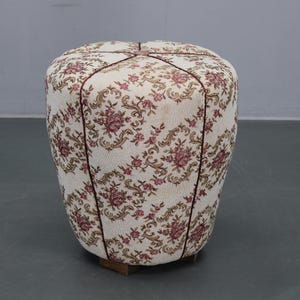 May include: An upholstered ottoman with a floral pattern in pink and brown on an off-white background. The ottoman has a rounded shape and is trimmed with a dark brown piping. It sits on a small wooden base.