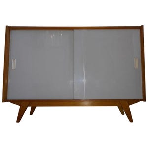 May include: A mid-century modern cabinet with a light wood frame and legs. The cabinet has two sliding doors with a frosted glass appearance and small, rectangular handles. The cabinet is approximately 48 inches wide.