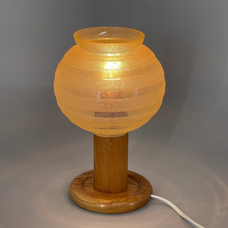 Puede incluir: Una l&aacute;mpara de mesa con una pantalla redonda de vidrio &aacute;mbar texturizado y una base de madera. La base tiene un tallo cil&iacute;ndrico y una plataforma redonda de madera.