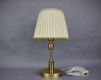 May include: A vintage brass table lamp with a pleated cream lampshade. The lamp has a round base and a slender stem. The lampshade is trimmed with a gold border. A white power cord with an on/off switch is visible.
