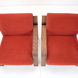 May include: Two mid-century modern armchairs. Each chair features a burnt orange seat and backrest, with a dark wood frame. The armrests are angled, and the chairs are set against a white background.
