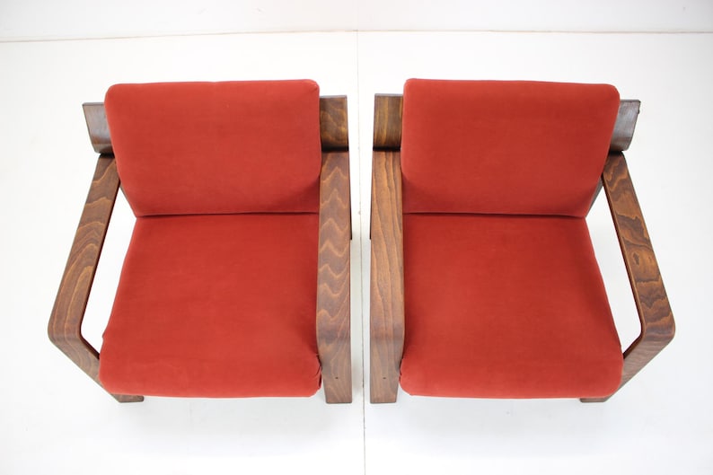 May include: Two mid-century modern armchairs. Each chair features a burnt orange seat and backrest, with a dark wood frame. The armrests are angled, and the chairs are set against a white background.