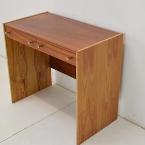 Peut inclure: Un bureau en bois avec deux tiroirs et des poignées argentées. Le bureau est en bois brun clair avec un grain de bois naturel.