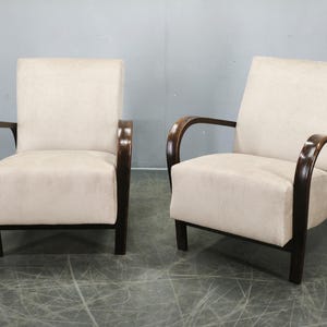 May include: A pair of armchairs featuring cream-coloured upholstered seats and backrests. The chairs have dark brown, curved wooden armrests and legs that extend from the base to the top of the armrest, providing a structured frame for the cushions.