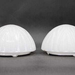 Puede incluir: Dos pantallas de lámpara de vidrio blanco en forma de cúpula. Cada pantalla tiene una serie de crestas verticales y curvas. Las pantallas tienen un acabado brillante y una base circular.