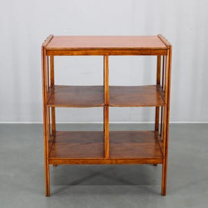 Puede incluir: Estantería de madera de tres niveles con acabado marrón cálido. Los estantes son rectangulares y están soportados por postes verticales. El estante superior tiene un borde ligeramente elevado. La unidad es independiente.