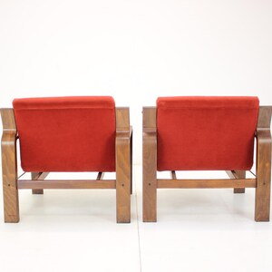 May include: Two mid-century modern armchairs with a wooden frame and a vibrant red velvet seat and back. The chairs are positioned side-by-side, showcasing their design and style. The wooden frames have a warm, natural finish.