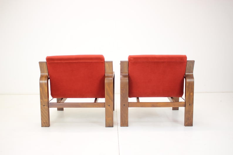 May include: Two mid-century modern armchairs with a wooden frame and a vibrant red velvet seat and back. The chairs are positioned side-by-side, showcasing their design and style. The wooden frames have a warm, natural finish.