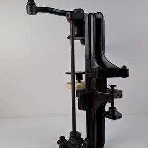 May include: A vintage black metal can sealer with a wooden handle. The device has a crank, a vertical rod, and various adjustable parts. It is designed to seal cans and bottles.