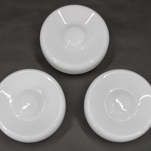 May include: Three white, round, ceramic bowls with a central recessed area. The bowls have a glossy finish and are arranged on a dark gray surface. The bowls are likely for serving or display.