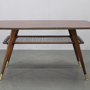 May include: Mid-century modern coffee table with a rectangular top and a lower shelf. The table features dark brown wooden legs with brass-colored tips. The top is a light brown color. The lower shelf has a woven design.