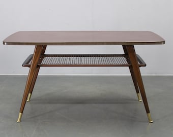 Puede incluir: Mesa de centro de mediados de siglo con tapa rectangular y estante inferior. La mesa tiene patas de madera marrón oscuro con puntas doradas. La tapa es de color marrón claro. El estante inferior tiene un diseño tejido.