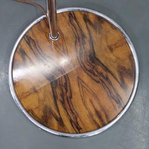 May include: Close-up of a floor lamp base. The base is a round, wooden disc with a polished finish, showcasing a natural wood grain pattern in shades of brown and black. A silver metal ring surrounds the wooden base.