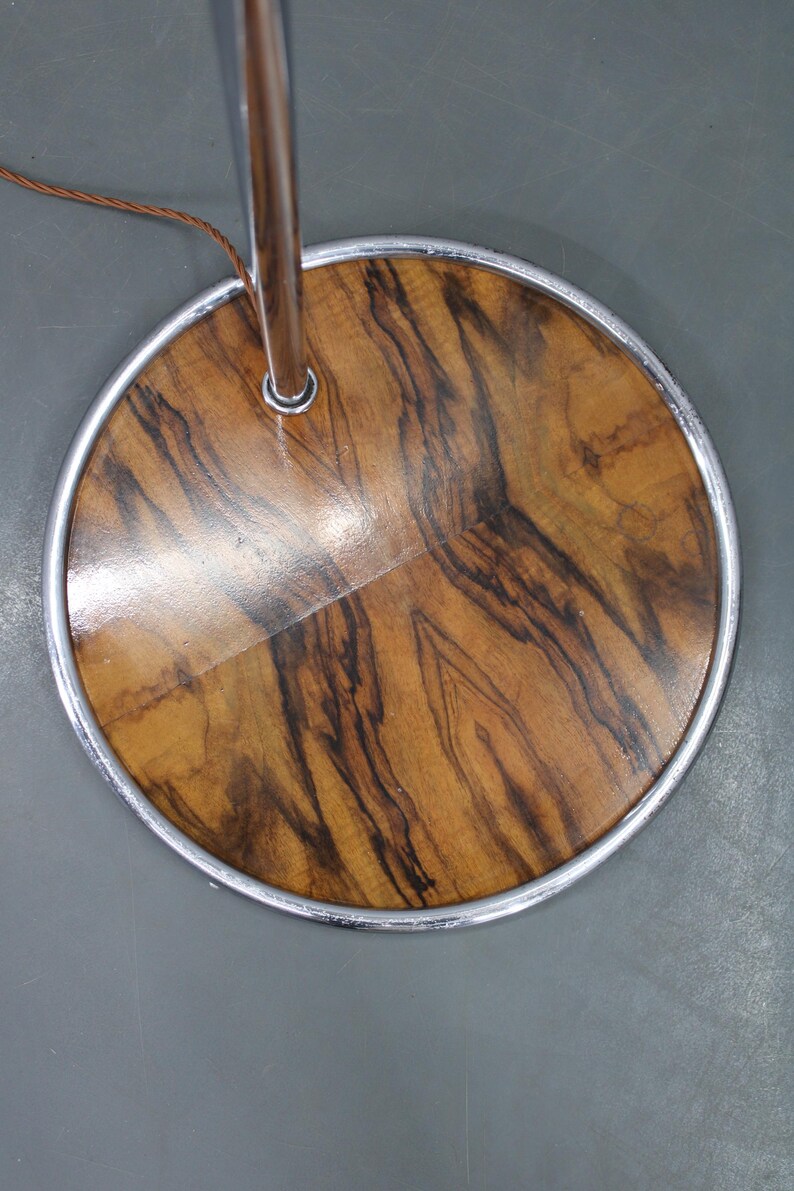 May include: Close-up of a floor lamp base. The base is a round, wooden disc with a polished finish, showcasing a natural wood grain pattern in shades of brown and black. A silver metal ring surrounds the wooden base.