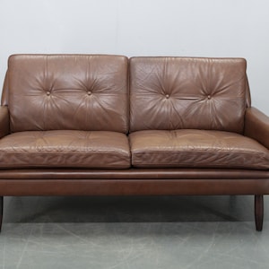 Puede incluir: Un sofá de cuero marrón de dos plazas con respaldo y cojines de asiento acolchados. El sofá tiene un perfil bajo con estructura y patas de madera. Los brazos están ligeramente curvados. Diseño moderno de mediados de siglo.