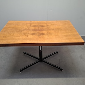May include: A square wooden table with a light brown surface and dark brown edges. The table is supported by a black, star-shaped metal base. The table appears to be approximately 30 inches wide.