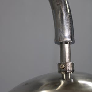 May include: Close-up of a modern lamp with a curved, brushed metal arm. The lamp features a white, rounded shade and a polished metal base. The design is minimalist and sleek, suitable for contemporary interiors.