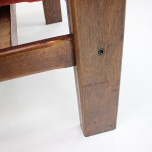 May include: Close-up of a wooden chair leg and frame. The wood has a warm, brown tone with visible grain and a smooth finish. A red fabric is visible on the seat. The chair appears to be vintage or antique.