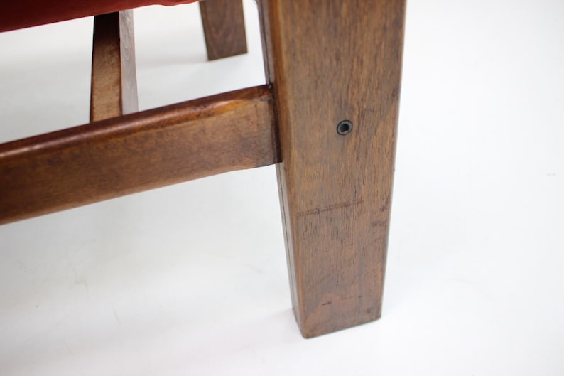 May include: Close-up of a wooden chair leg and frame. The wood has a warm, brown tone with visible grain and a smooth finish. A red fabric is visible on the seat. The chair appears to be vintage or antique.
