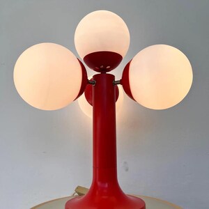 May include: A red and white table lamp with four spherical light bulbs. The lamp has a red base and stem, with the bulbs arranged around the top. The bulbs are white and emit a soft glow, creating a retro aesthetic.