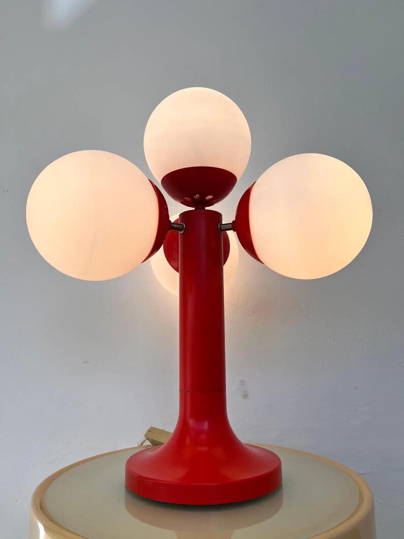 May include: A red and white table lamp with four spherical light bulbs. The lamp has a red base and stem, with the bulbs arranged around the top. The bulbs are white and emit a soft glow, creating a retro aesthetic.