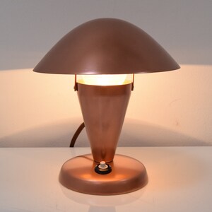 May include: A copper-colored desk lamp with a mushroom-shaped shade and a conical base. The lamp is turned on and illuminating the room.