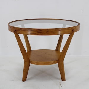 May include: A round wooden side table with a glass top and a lower shelf. The table has a light brown wood finish and three legs.