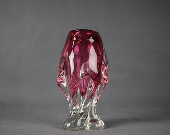 Puede incluir: Un jarrón de vidrio rosa con una forma abstracta y orgánica. El jarrón tiene una base de vidrio transparente y un cuerpo de vidrio rosa con un diseño en espiral.