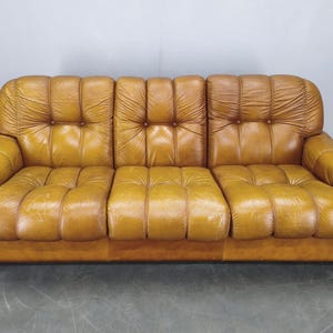 Könnte beinhalten: Ein Vintage-Sofa mit drei Sitzplätzen in warmem, goldbraunem Leder. Das Sofa hat ein getuftetes Design auf den Kissen und der Rückenlehne. Die Armlehnen sind abgerundet, und das Sofa ist niedrig. Wohnzimmermöbel.
