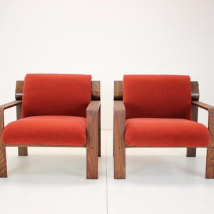 May include: Two mid-century modern armchairs. Each chair has a wooden frame and legs, with a rectangular seat and backrest upholstered in a burnt orange fabric. The chairs are set against a white background.