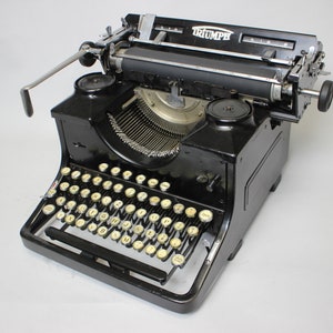 Puede incluir: Una máquina de escribir Triumph negra vintage con teclas color crema. La máquina de escribir tiene una palanca de retorno de carro plateada y una guía de papel. La palabra "TRIUMPH" es visible en la parte superior. Un clásico de los suministros de oficina.