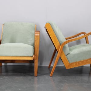 May include: Two mid-century modern armchairs. The chairs have light green upholstery and wooden frames. The wooden frames have a natural finish. One chair is upright, and the other is angled.