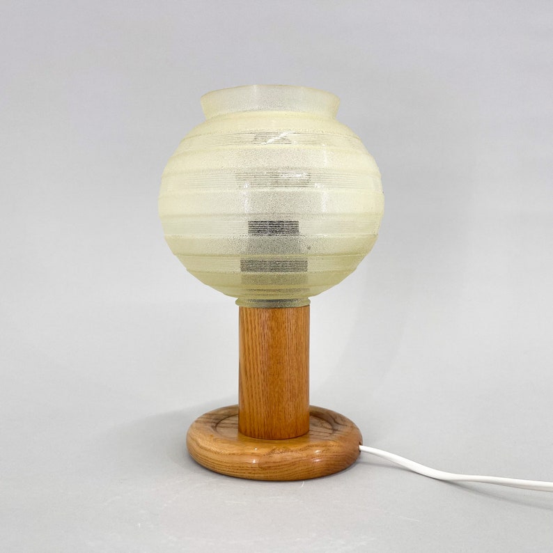 Puede incluir: Una l&aacute;mpara de mesa con una pantalla blanca texturizada en forma de globo y una base de madera. La base tiene una columna cil&iacute;ndrica y una base redonda y plana.