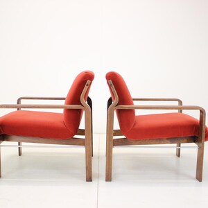 May include: Two mid-century modern armchairs with a curved design. The chairs have a wooden frame and legs, with a vibrant red upholstered seat and back. The armrests are also made of wood. The chairs are set against a white background.