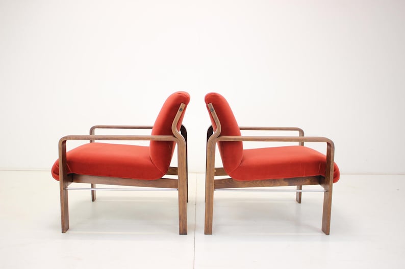 May include: Two mid-century modern armchairs with a curved design. The chairs have a wooden frame and legs, with a vibrant red upholstered seat and back. The armrests are also made of wood. The chairs are set against a white background.