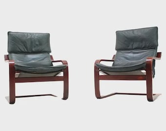 May include: Two mid-century modern armchairs with dark green leather cushions and backs. The chairs have a dark wood frame with curved armrests and a low profile. The chairs are set against a white background.