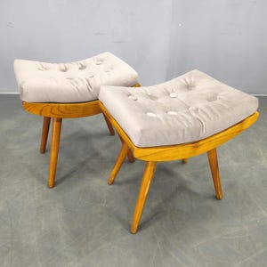 Puede incluir: Dos otomanas de mediados de siglo. Cada una tiene un cojín beige claro con botones sobre un marco de madera curvado de color marrón claro. Las otomanas tienen tres patas anguladas y cónicas de madera más clara.