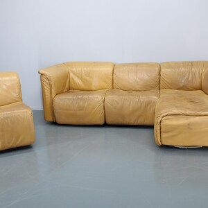 May include: A large, tan leather sectional sofa with a matching armchair. The sofa has a chaise lounge on the right side and is made of multiple pieces that can be arranged in different configurations.