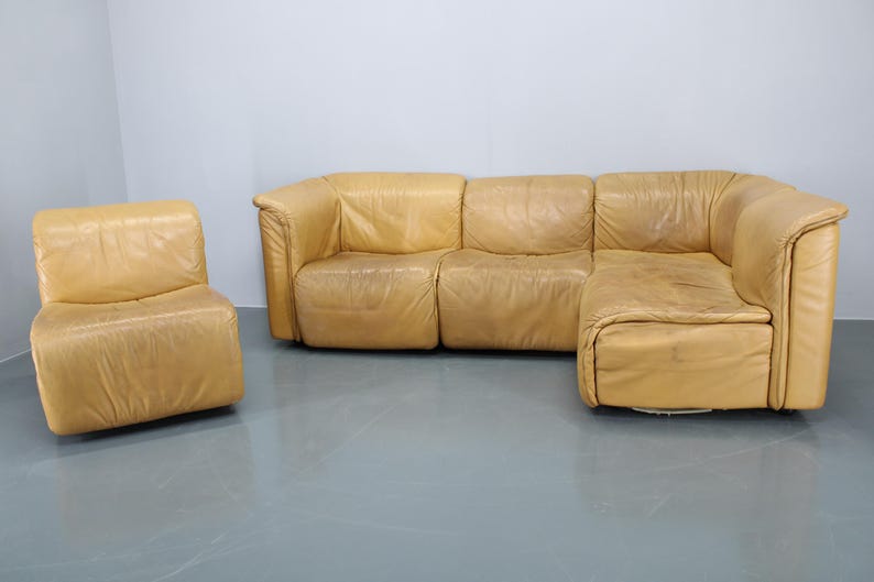 May include: A large, tan leather sectional sofa with a matching armchair. The sofa has a chaise lounge on the right side and is made of multiple pieces that can be arranged in different configurations.
