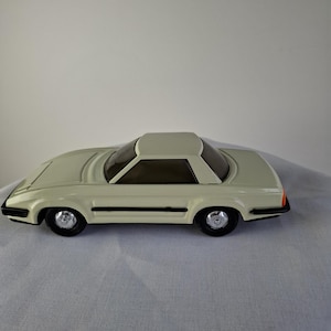 Pode incluir: Um carro de brinquedo creme com capota rígida. O carro tem detalhes em preto, pneus pretos e uma pequena luz laranja na traseira. É um modelo vintage, provavelmente dos anos 70 ou 80.