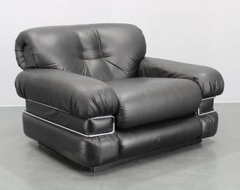 May include: A black leather armchair with a rounded design. The chair has a square base and thick, cushioned arms. White trim accents the edges of the base and arms. The chair is a modern style.