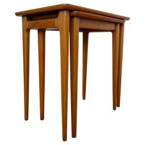 May include: A set of two nesting tables crafted from light brown wood. Each table features a rectangular top with rounded edges and four slender, tapered legs that extend outward from the frame, providing a stable and elevated structure.