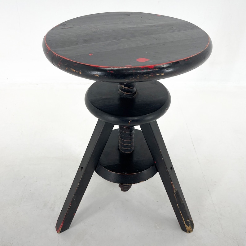 May include: A black wooden adjustable stool with three legs and a round seat. The stool has a red painted edge and a worn finish.