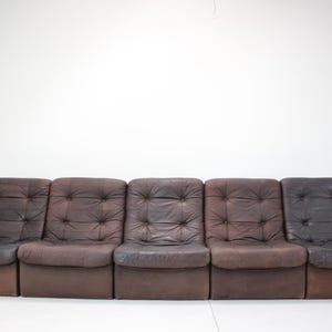 May include: A brown leather sofa with five individual sections. Each section has a tufted back and seat. The sofa has a low profile and is set against a white wall.