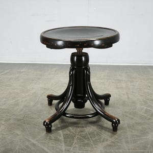 May include: A black wooden stool with a round seat and a central adjustable screw mechanism. The stool features four curved legs connected by a circular support ring near the base. The finish shows signs of wear with visible scuffs and wood grain.