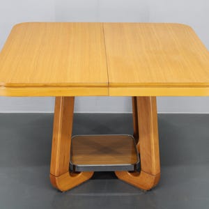 May include: A light-colored wooden table with a square top and rounded corners. The table has a unique base with a shelf and a light-colored finish. The table's dimensions are approximately 30 inches in length and width.