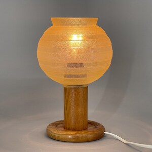 Puede incluir: Una l&aacute;mpara de mesa con una pantalla redonda de vidrio naranja claro texturizado y una base de madera. La base es un cilindro con una plataforma redonda de madera.